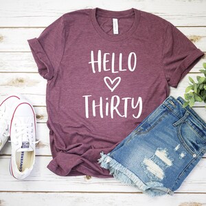 May include: A maroon t-shirt with the text "Hello Thirty" in white with a white heart between the words.