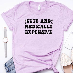 May include: A light purple t-shirt with the text "CUTE AND MEDICALLY EXPENSIVE" in black. The shirt is styled with a watch, jeans, a phone, and shoes.