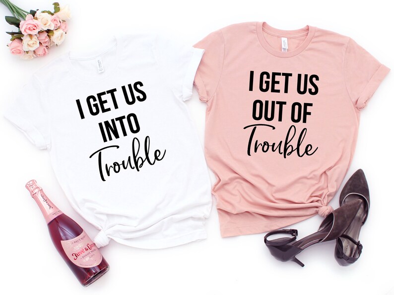 Op de afbeelding: Twee T-shirts met bijpassende tekst. Een wit T-shirt zegt "I get us into trouble" en een roze T-shirt zegt "I get us out of trouble".