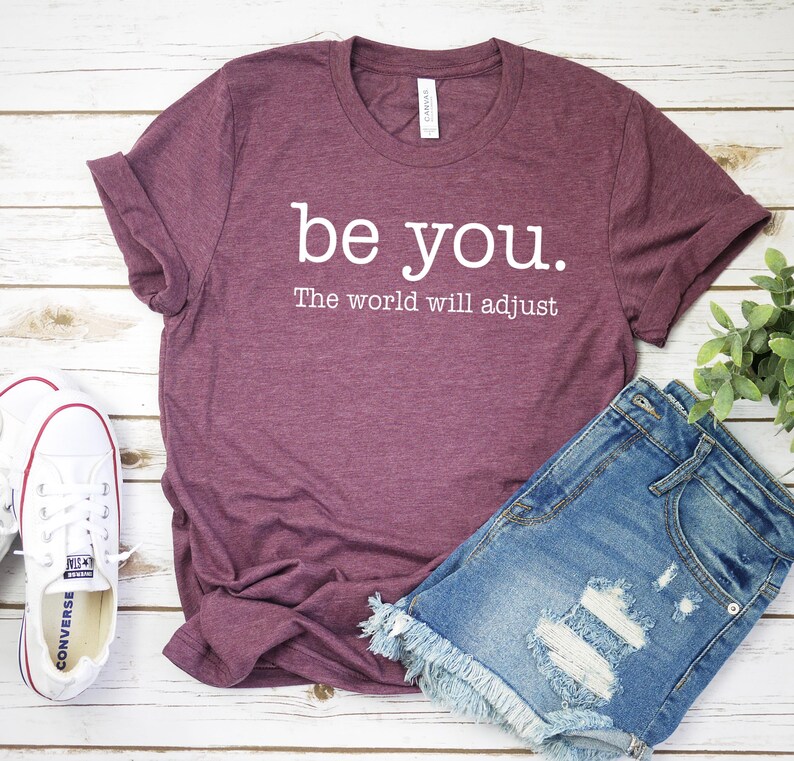Puede incluir: Una camiseta granate con el texto blanco "be you. The world will adjust". Se combina con pantalones cortos de mezclilla y zapatillas blancas sobre una superficie de madera blanca. Una planta verde est&aacute; en el fondo.