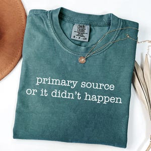Puede incluir: Una camiseta de manga corta color verde azulado con cuello redondo que presenta el texto blanco "primary source or it didn't happen" impreso en el frente. La camiseta tiene una etiqueta de la marca Comfort Colors en el cuello interior indicando la talla S.