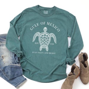 Op de afbeelding: Teal langarm T-shirt met een witte afbeelding van een zeeschildpad en de tekst "GULF OF MEXICO EST. 1550 STAY SALTY AND RESIST".