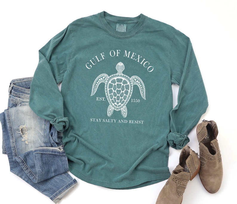 Op de afbeelding: Teal langarm T-shirt met een witte afbeelding van een zeeschildpad en de tekst "GULF OF MEXICO EST. 1550 STAY SALTY AND RESIST".