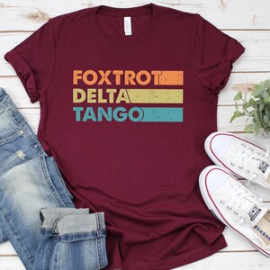 以下が含まれることがあります： ワインレッドのTシャツに、ヴィンテージスタイルで「FOXTROT DELTA TANGO」という文字がカラフルなストライプでプリントされています。