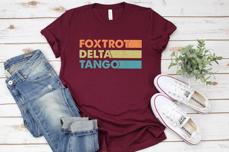 以下が含まれることがあります： ワインレッドのTシャツに、ヴィンテージスタイルで「FOXTROT DELTA TANGO」という文字がカラフルなストライプでプリントされています。