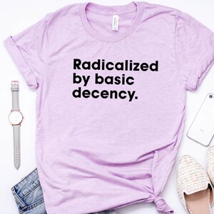 May include: A light purple t-shirt with the text "Radicalized by basic decency." is displayed on a white surface. A gray watch, a pair of jeans, a smartphone, and a pair of cream-colored shoes are also visible.