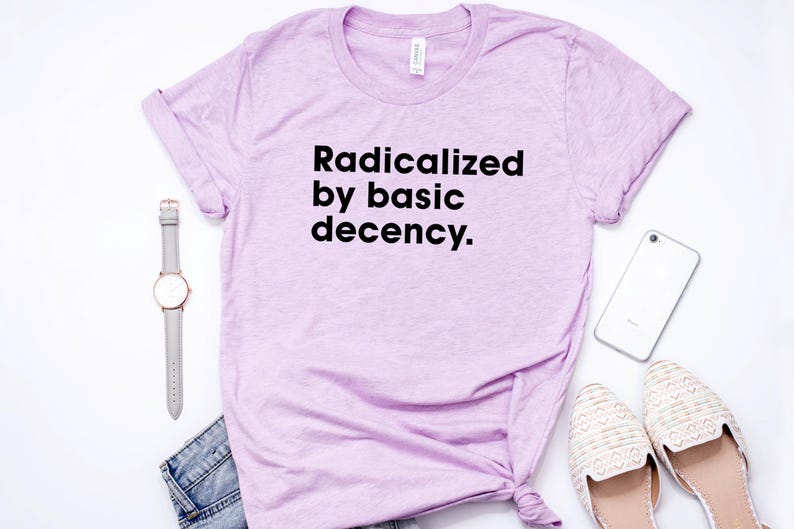 May include: A light purple t-shirt with the text "Radicalized by basic decency." is displayed on a white surface. A gray watch, a pair of jeans, a smartphone, and a pair of cream-colored shoes are also visible.