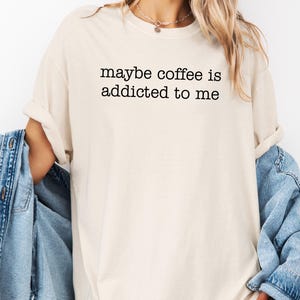 May include: Cream-colored t-shirt with the text "maybe coffee is addicted to me" in black. The shirt has a crew neck and short sleeves. A light blue denim jacket is draped over the shoulders.