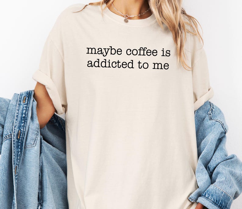 May include: Cream-colored t-shirt with the text "maybe coffee is addicted to me" in black. The shirt has a crew neck and short sleeves. A light blue denim jacket is draped over the shoulders.