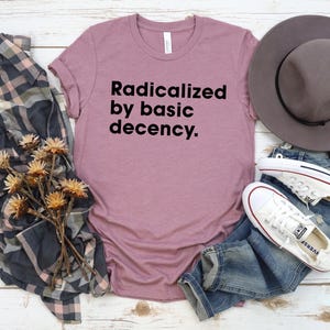 May include: Mauve t-shirt with the text "Radicalized by basic decency." A gray hat, blue jeans, and white sneakers are arranged with a plaid shirt and dried flowers on a white wood surface. Casual wear.