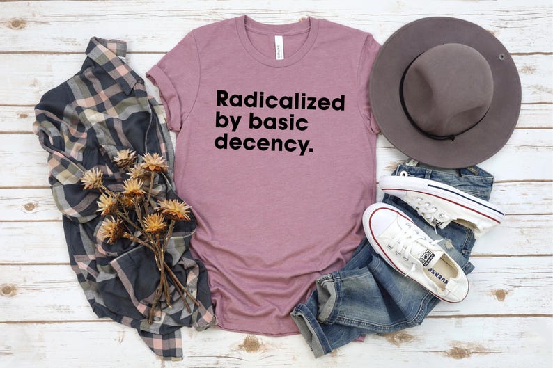 May include: Mauve t-shirt with the text "Radicalized by basic decency." A gray hat, blue jeans, and white sneakers are arranged with a plaid shirt and dried flowers on a white wood surface. Casual wear.