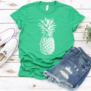 Pode incluir: Uma camiseta verde com um gr&aacute;fico de abacaxi branco. A camiseta &eacute; combinada com shorts jeans e t&ecirc;nis brancos.