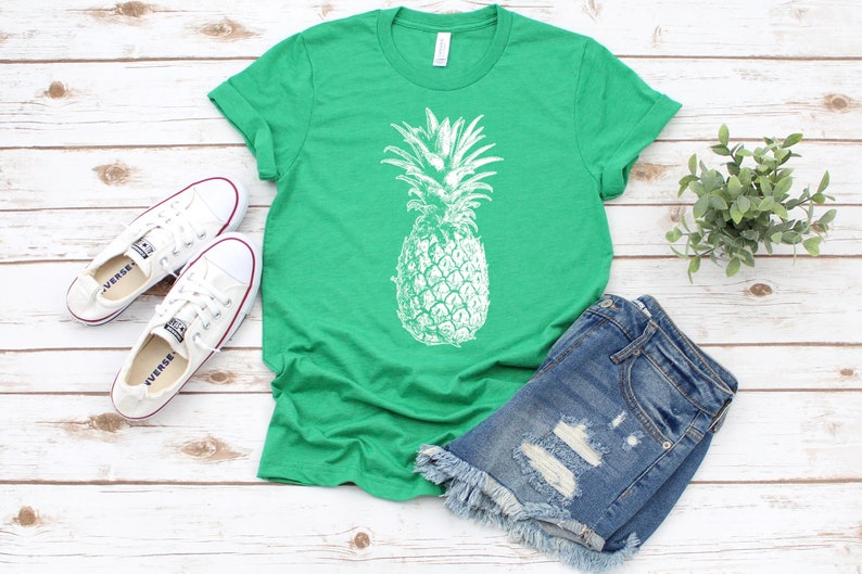 Pode incluir: Uma camiseta verde com um gr&aacute;fico de abacaxi branco. A camiseta &eacute; combinada com shorts jeans e t&ecirc;nis brancos.