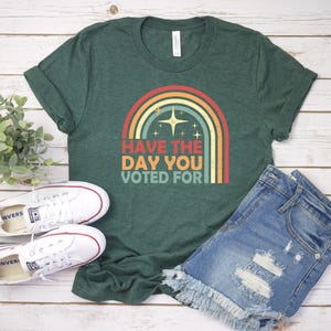 May include: Green t-shirt with a rainbow graphic and the text "Have the day you voted for." The shirt is paired with denim shorts and white sneakers. The image is on a light wood background.