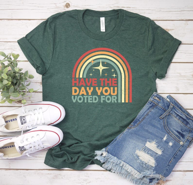 May include: Green t-shirt with a rainbow graphic and the text "Have the day you voted for." The shirt is paired with denim shorts and white sneakers. The image is on a light wood background.