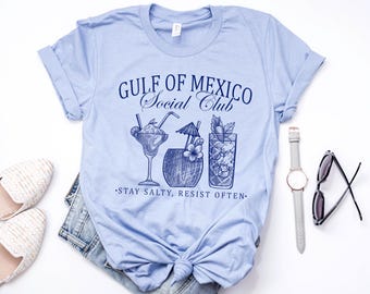 Puede incluir: Camiseta azul claro con el texto "Gulf of Mexico Social Club" y tres ilustraciones de cócteles. El texto debajo de las ilustraciones dice "Stay Salty, Resist Often."