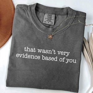 May include: A charcoal gray Comfort Colors t-shirt with the phrase "that wasn't very evidence based of you" in white. A gold pendant necklace and a brown hat are also visible.