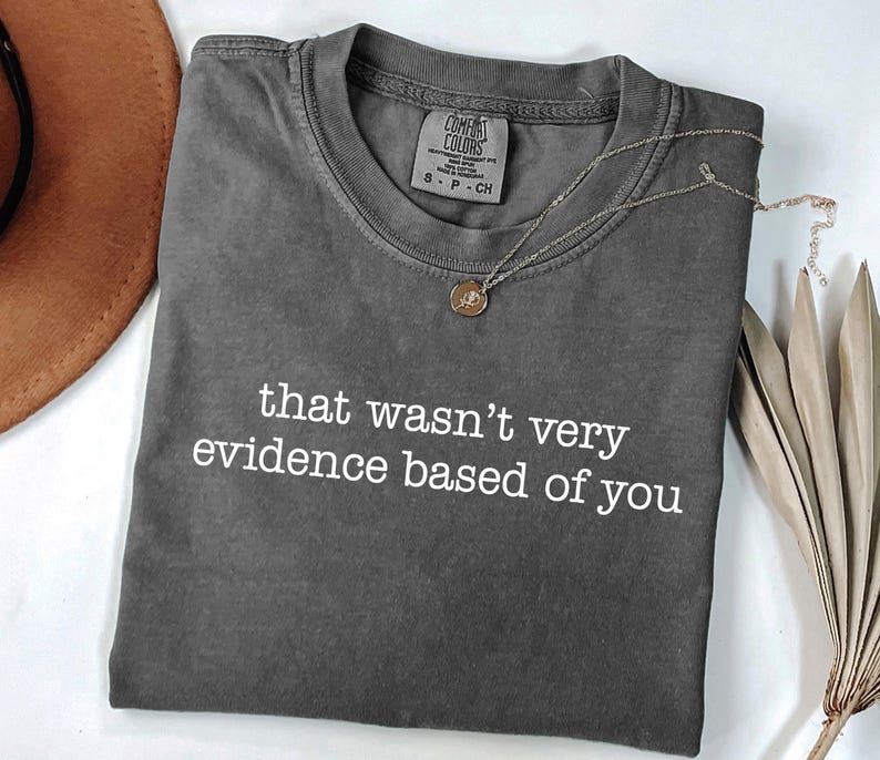May include: A charcoal gray Comfort Colors t-shirt with the phrase "that wasn't very evidence based of you" in white. A gold pendant necklace and a brown hat are also visible.
