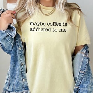 May include: A light yellow t-shirt with the text "maybe coffee is addicted to me" in black. The shirt is paired with a blue denim jacket. A gold necklace is visible.