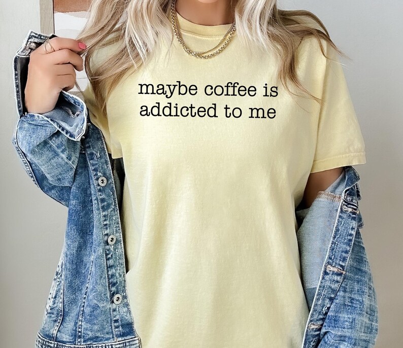 May include: A light yellow t-shirt with the text "maybe coffee is addicted to me" in black. The shirt is paired with a blue denim jacket. A gold necklace is visible.