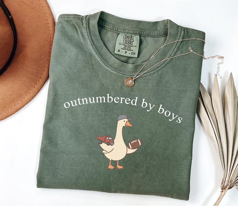 May include: An olive green Comfort Colors t-shirt with the white text "outnumbered by boys". The shirt features a cartoon goose wearing a hat and holding a football and a red truck. A gold necklace is draped across the shirt.