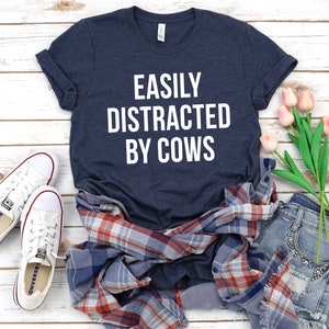 May include: A navy blue t-shirt with the text "EASILY DISTRACTED BY COWS" in white lettering. The shirt is laid out on a white wooden surface with a plaid shirt, denim shorts, and pink flowers.