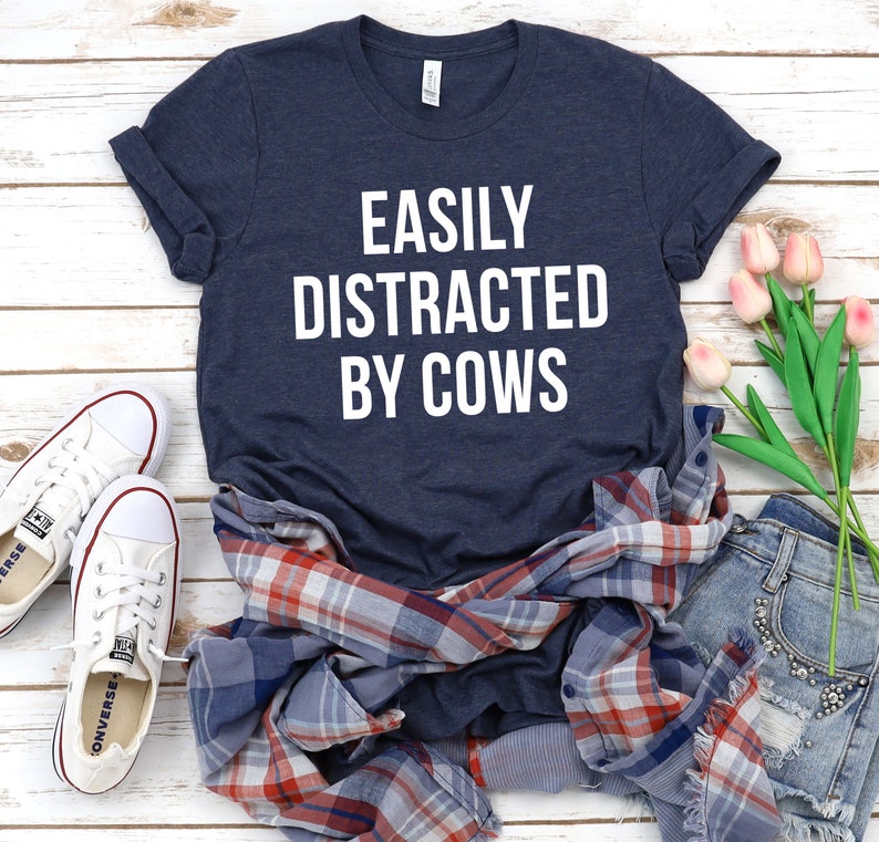 May include: A navy blue t-shirt with the text "EASILY DISTRACTED BY COWS" in white lettering. The shirt is laid out on a white wooden surface with a plaid shirt, denim shorts, and pink flowers.