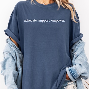 Op de afbeelding: Een marineblauw t-shirt met de woorden "advocate. support. empower." in het wit gedrukt. Het shirt heeft een losse pasvorm en opgerolde mouwen. Een lichtblauwe spijkerjas wordt over het shirt gedragen.