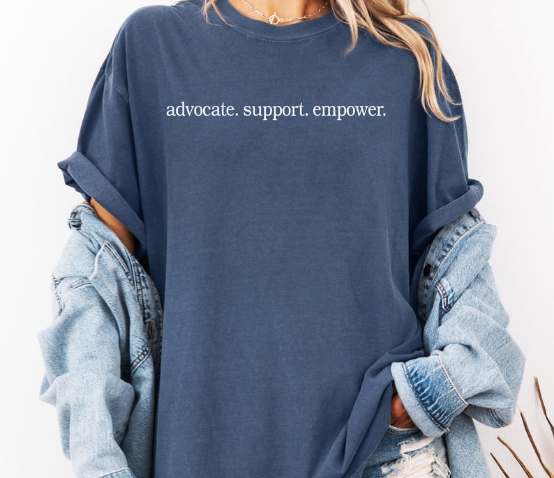 Op de afbeelding: Een marineblauw t-shirt met de woorden "advocate. support. empower." in het wit gedrukt. Het shirt heeft een losse pasvorm en opgerolde mouwen. Een lichtblauwe spijkerjas wordt over het shirt gedragen.