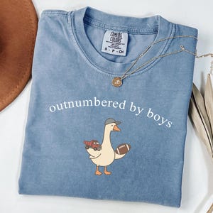 May include: A blue Comfort Colors t-shirt with the phrase "outnumbered by boys" printed in white. The shirt features a cartoon goose wearing a baseball cap and holding a football and a toy truck. A gold necklace with a pendant is draped across the shirt.