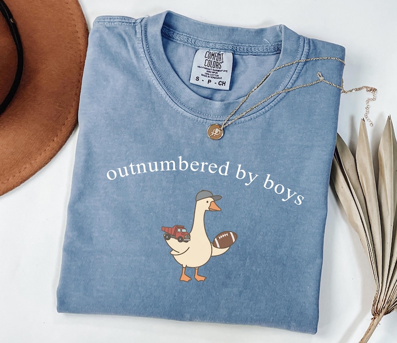 May include: A blue Comfort Colors t-shirt with the phrase "outnumbered by boys" printed in white. The shirt features a cartoon goose wearing a baseball cap and holding a football and a toy truck. A gold necklace with a pendant is draped across the shirt.
