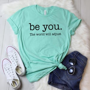 Puede incluir: Una camiseta verde menta con el texto "be you. The world will adjust" en negro. La camiseta se combina con pantalones cortos de mezclilla azules, zapatillas blancas y gafas de sol. Los art&iacute;culos est&aacute;n colocados sobre una superficie blanca y esponjosa.