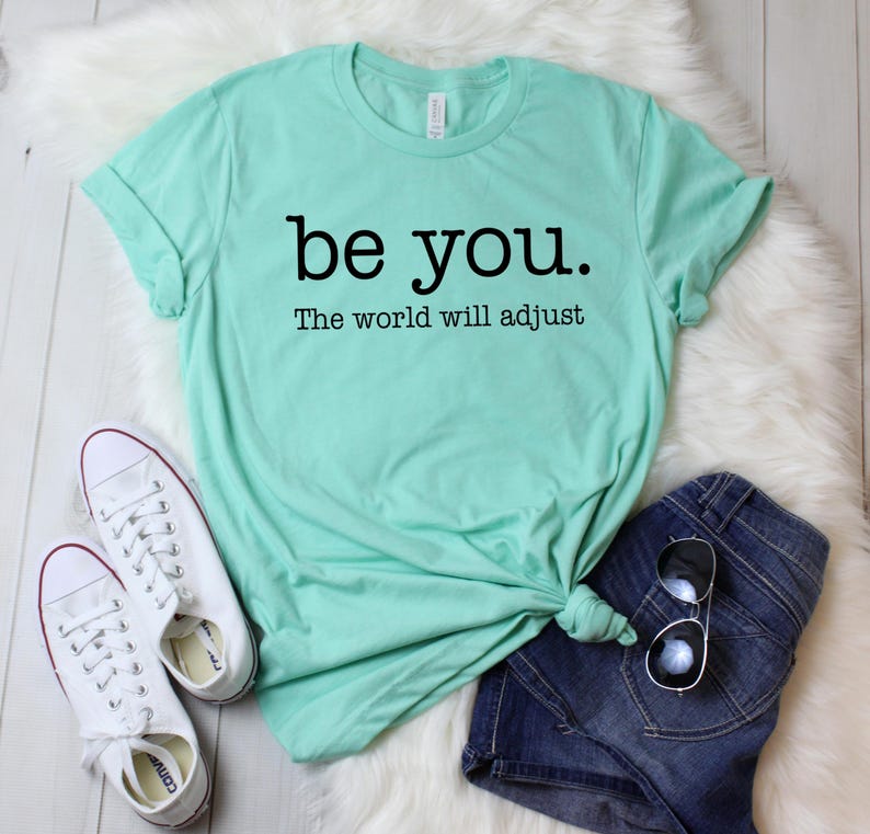 Puede incluir: Una camiseta verde menta con el texto "be you. The world will adjust" en negro. La camiseta se combina con pantalones cortos de mezclilla azules, zapatillas blancas y gafas de sol. Los art&iacute;culos est&aacute;n colocados sobre una superficie blanca y esponjosa.