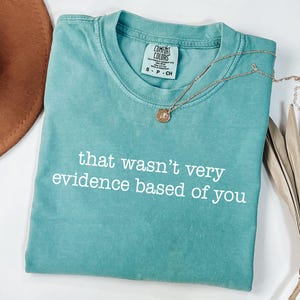 May include: A teal Comfort Colors t-shirt with the phrase "that wasn't very evidence based of you" in white text. A gold necklace with a pendant rests on the shirt. A brown hat and dried plant are in the background.