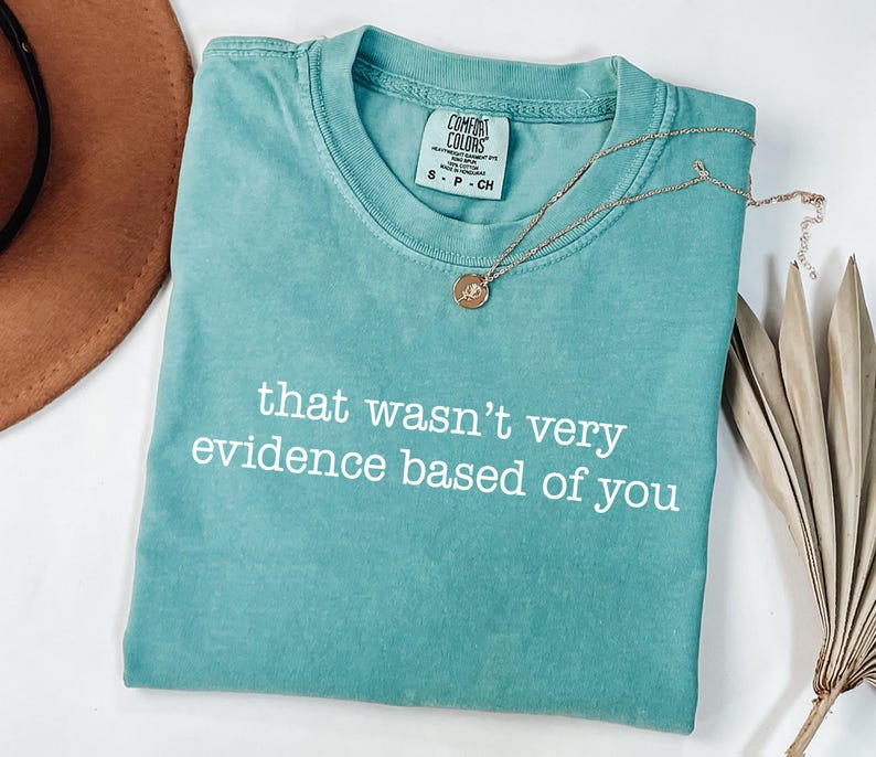 May include: A teal Comfort Colors t-shirt with the phrase "that wasn't very evidence based of you" in white text. A gold necklace with a pendant rests on the shirt. A brown hat and dried plant are in the background.