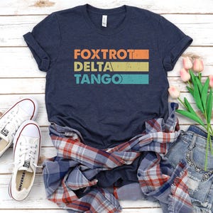 以下が含まれることがあります： 「FOXTROT DELTA TANGO」とヴィンテージスタイルのグラフィックがプリントされたネイビーブルーのTシャツ。シャツは折り畳まれ、木製の表面に、チェックシャツ、デニムのショートパンツ、花と一緒にディスプレイされています。