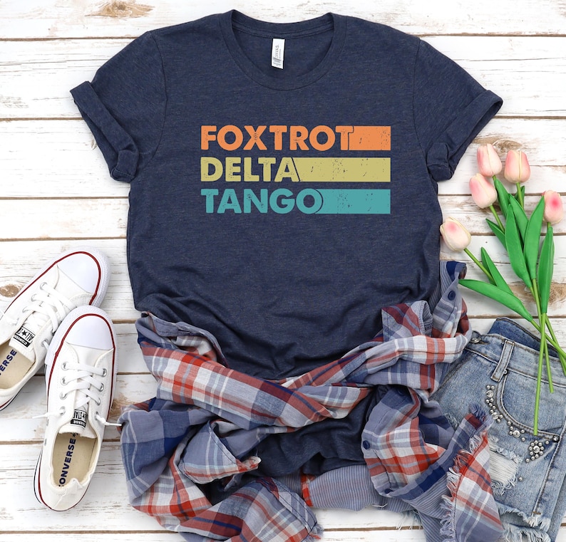以下が含まれることがあります： 「FOXTROT DELTA TANGO」とヴィンテージスタイルのグラフィックがプリントされたネイビーブルーのTシャツ。シャツは折り畳まれ、木製の表面に、チェックシャツ、デニムのショートパンツ、花と一緒にディスプレイされています。