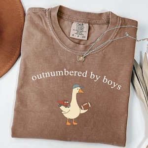 May include: A brown short-sleeve t-shirt with a crew neckline featuring the white text "outnumbered by boys" above a graphic of a goose wearing a grey baseball cap, holding a red toy dump truck and a brown football, with a Comfort Colors label.