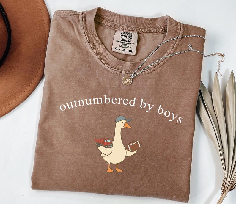 May include: A brown short-sleeve t-shirt with a crew neckline featuring the white text "outnumbered by boys" above a graphic of a goose wearing a grey baseball cap, holding a red toy dump truck and a brown football, with a Comfort Colors label.