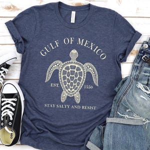 Puede incluir: Una camiseta azul marino con un gr&aacute;fico blanco de una tortuga y el texto "GULF OF MEXICO" y "STAY SALTY AND RESIST". La camiseta se combina con vaqueros azules y zapatillas negras.