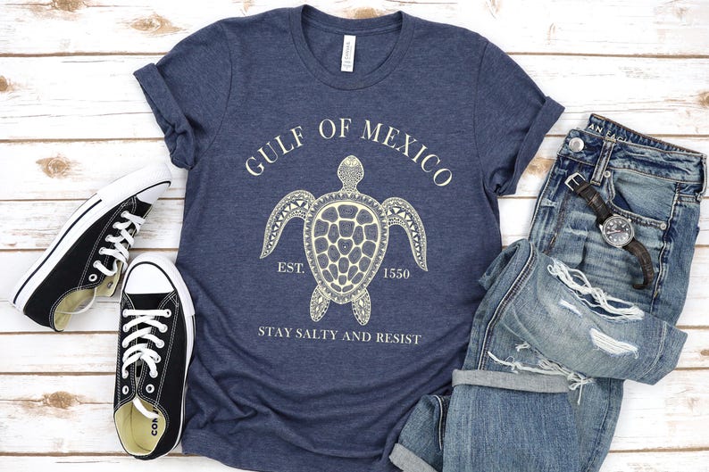 Puede incluir: Una camiseta azul marino con un gr&aacute;fico blanco de una tortuga y el texto "GULF OF MEXICO" y "STAY SALTY AND RESIST". La camiseta se combina con vaqueros azules y zapatillas negras.