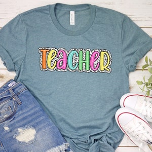 Puede incluir: Una camiseta azul turquesa con la palabra "teacher" en letras de colores con un patr&oacute;n de lunares blancos. La camiseta est&aacute; sobre una superficie de madera blanca con un par de zapatillas blancas y un par de pantalones cortos de mezclilla azul.