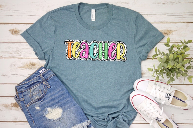 Puede incluir: Una camiseta azul turquesa con la palabra "teacher" en letras de colores con un patr&oacute;n de lunares blancos. La camiseta est&aacute; sobre una superficie de madera blanca con un par de zapatillas blancas y un par de pantalones cortos de mezclilla azul.
