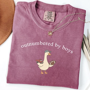May include: A mauve Comfort Colors t-shirt with the phrase "outnumbered by boys" printed in white. The shirt features a cartoon goose wearing a cap and holding a football and a toy truck. A gold necklace and a brown hat are also visible.