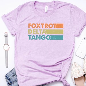 以下が含まれることがあります： 薄い紫色のTシャツに、ヴィンテージスタイルのグラフィックがプリントされています。グラフィックには「FOXTROT DELTA TANGO」という文字がオレンジ、イエロー、ターコイズで書かれています。