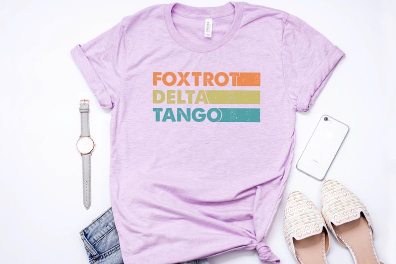 以下が含まれることがあります： 薄い紫色のTシャツに、ヴィンテージスタイルのグラフィックがプリントされています。グラフィックには「FOXTROT DELTA TANGO」という文字がオレンジ、イエロー、ターコイズで書かれています。