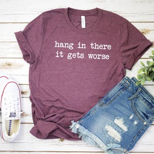 May include: A maroon t-shirt with the text "hang in there it gets worse" in white. The shirt is paired with denim shorts and white Converse sneakers. The items are arranged on a light wood-grain surface.