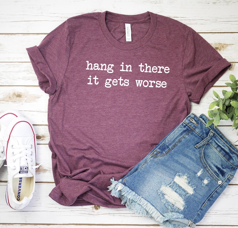 May include: A maroon t-shirt with the text "hang in there it gets worse" in white. The shirt is paired with denim shorts and white Converse sneakers. The items are arranged on a light wood-grain surface.