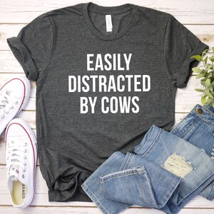 May include: A dark gray t-shirt with the text "EASILY DISTRACTED BY COWS" printed in white on the front. The shirt is laid flat on a white wooden surface with a pair of white sneakers and blue jeans.