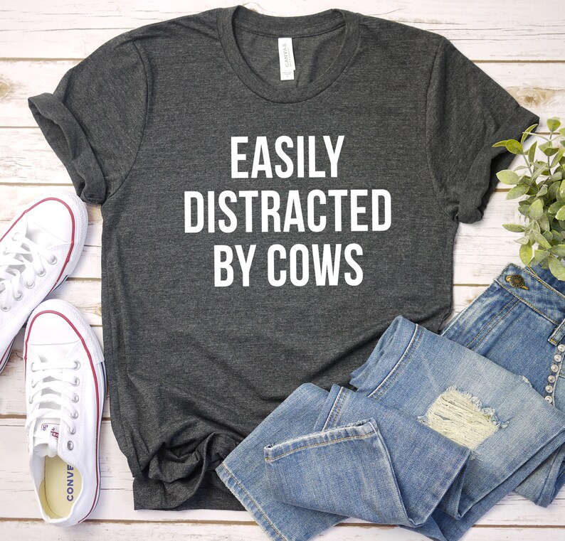 May include: A dark gray t-shirt with the text "EASILY DISTRACTED BY COWS" printed in white on the front. The shirt is laid flat on a white wooden surface with a pair of white sneakers and blue jeans.